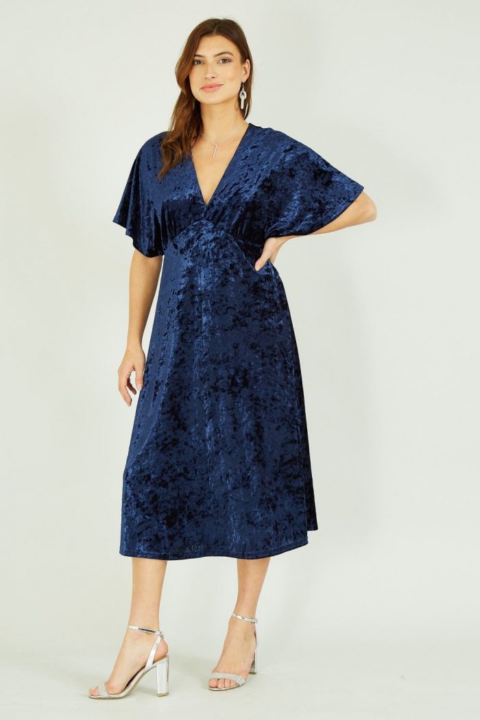 woman wearing a velvet navy dress with a plunging v-shaped neck like, short pleated sleeves and a skirt that falls below the knee